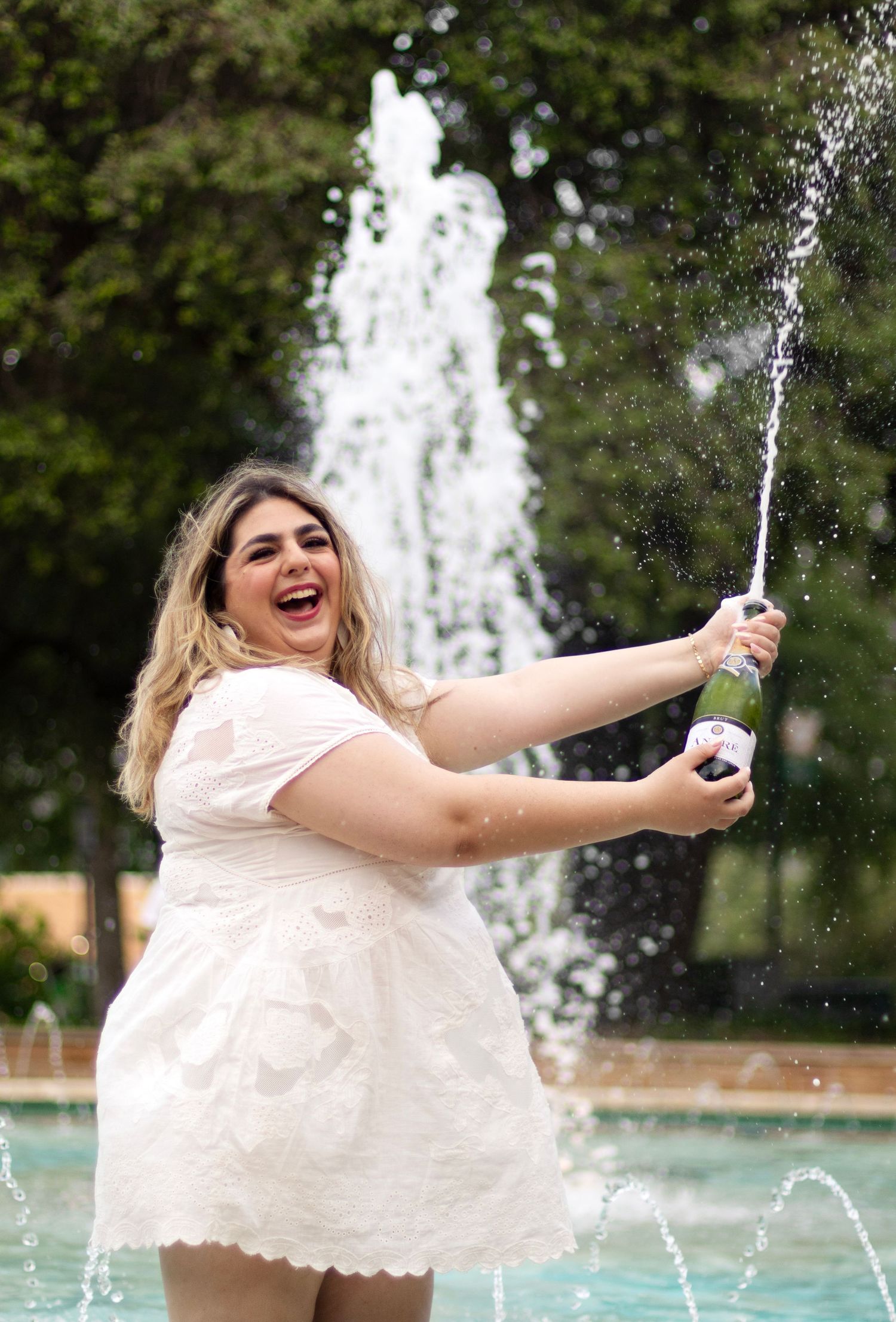 A college student pops a champagne in front of a fountain on campus.