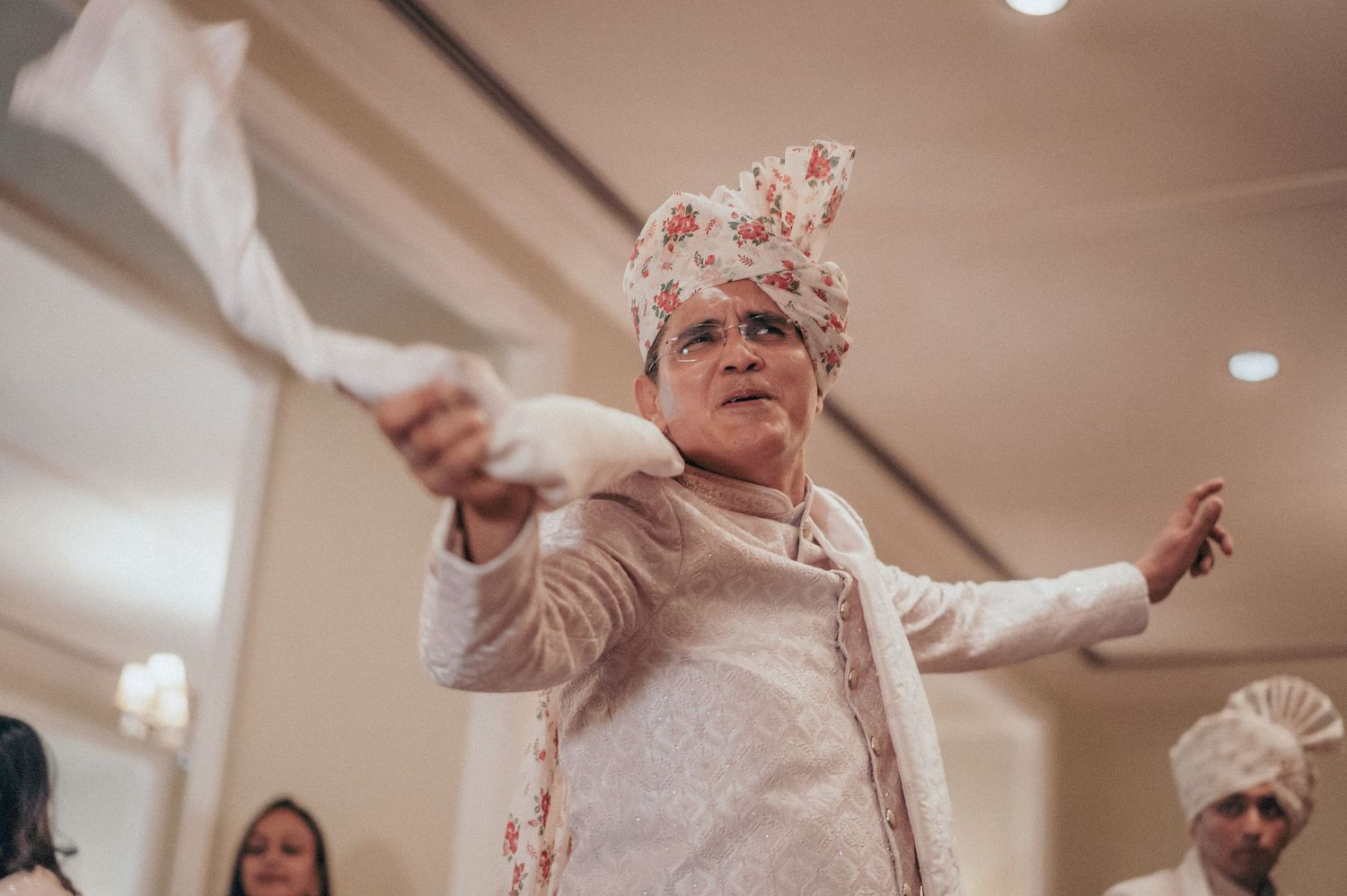 Elderly man in floral turban and white sherwani dancing energetically at Indian wedding celebration.