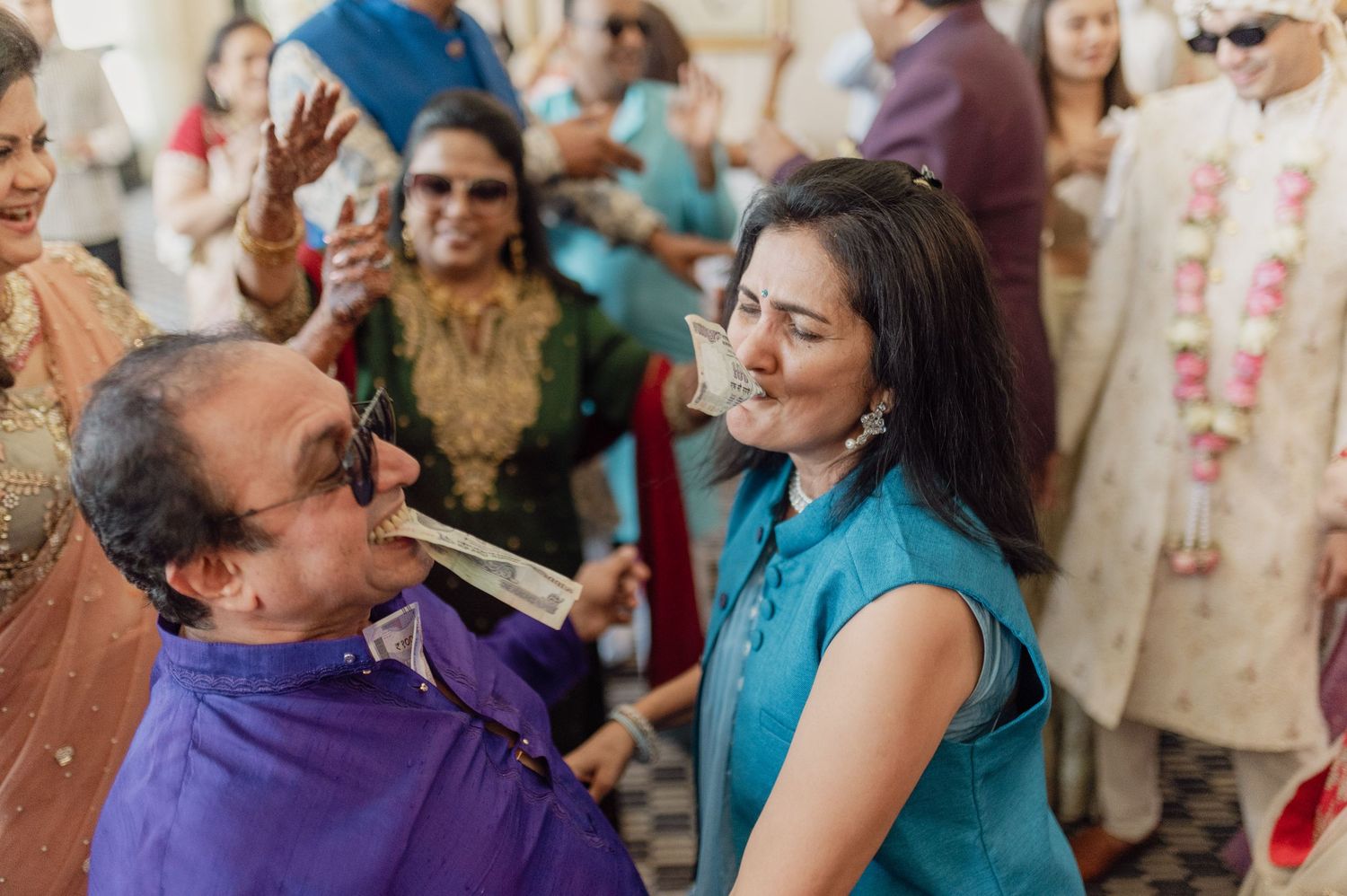 Man in purple playfully drinks from bottle held by smiling woman in teal at vibrant Indian wedding celebration.