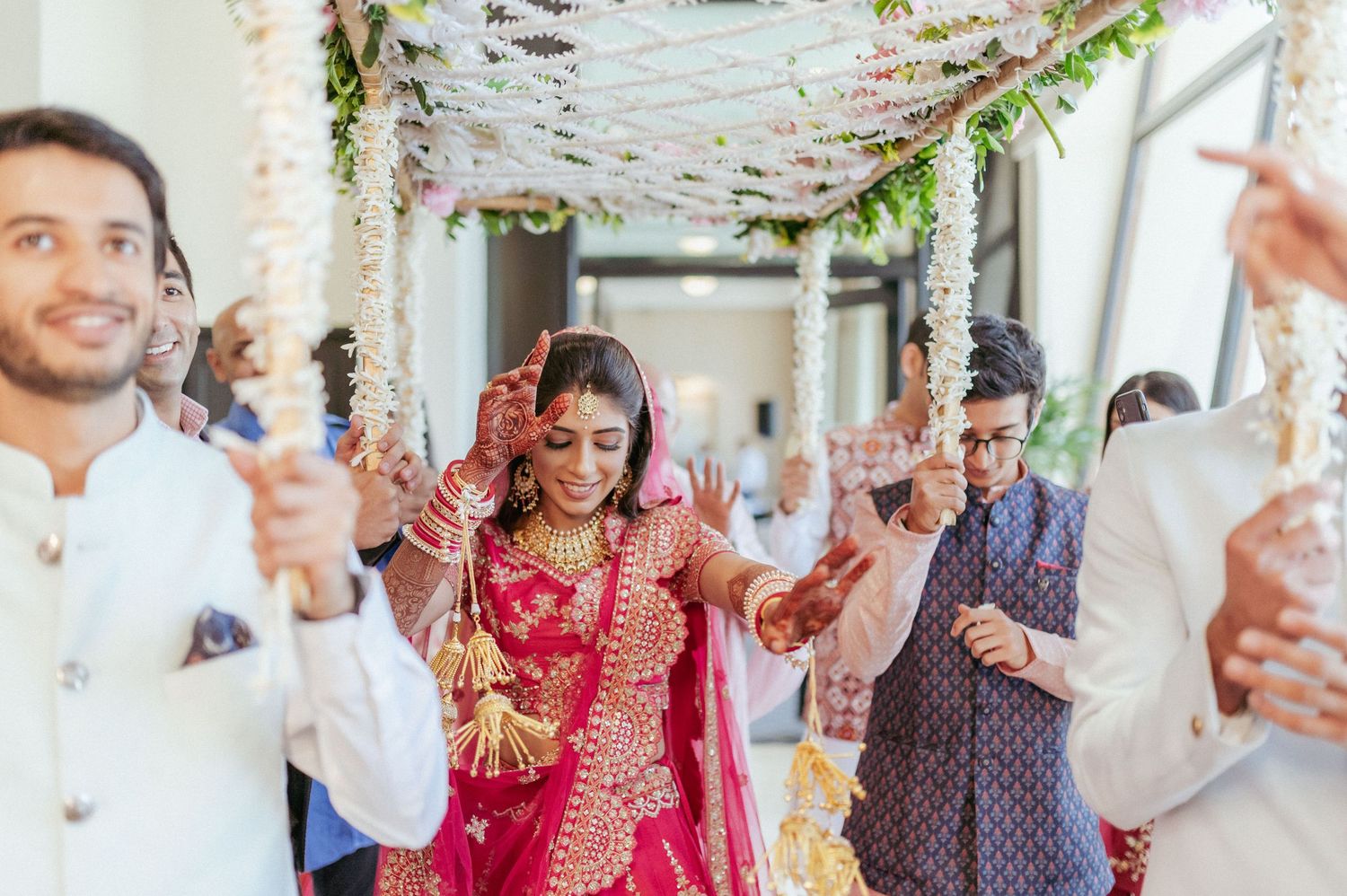 Bride in red bridal lehenga walks gracefully under pink floral canopy at Indian wedding.