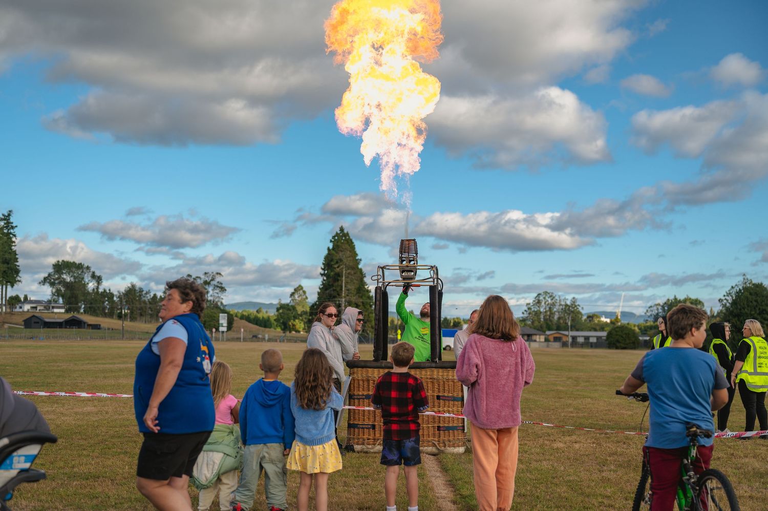 Crowd watches a hot air balloon burner shoot flames into the sky at an outdoor festival event.