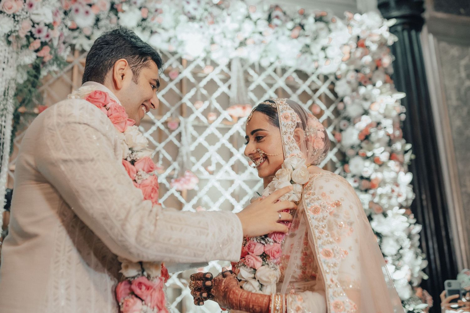 Indian bride and groom smiling warmly during jaal exchange ceremony with floral wedding backdrop.
