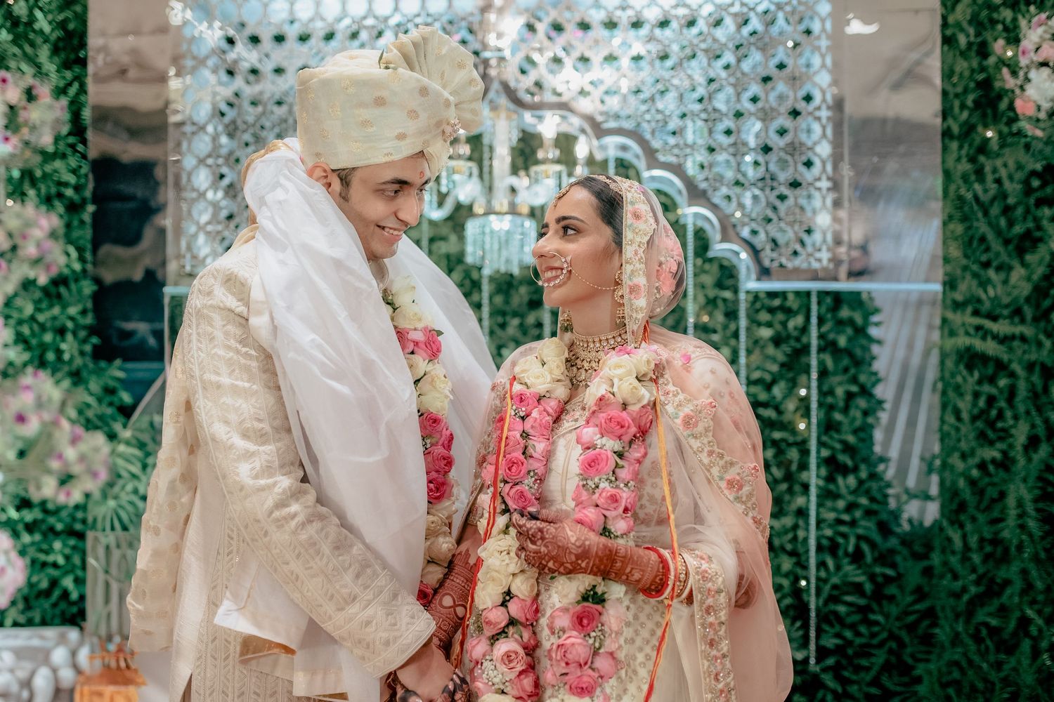Indian bride and groom in traditional wedding attire with floral garlands smiling at each other during ceremony.