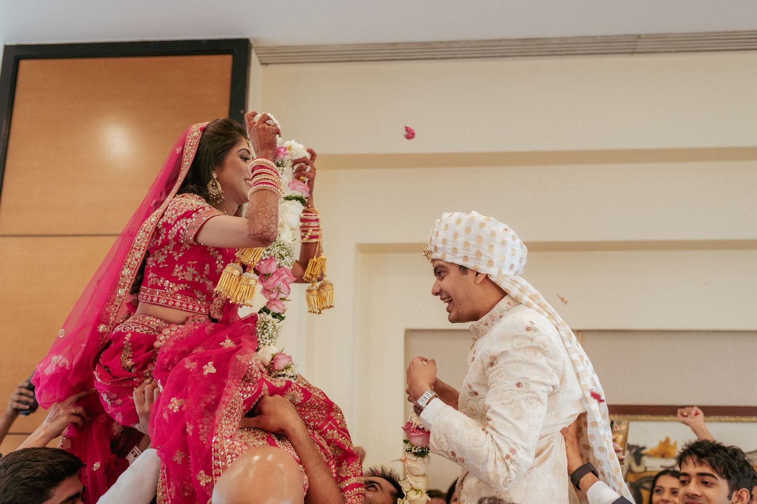 Indian bride in red lehenga and groom in white sherwani exchange garlands at wedding ceremony.