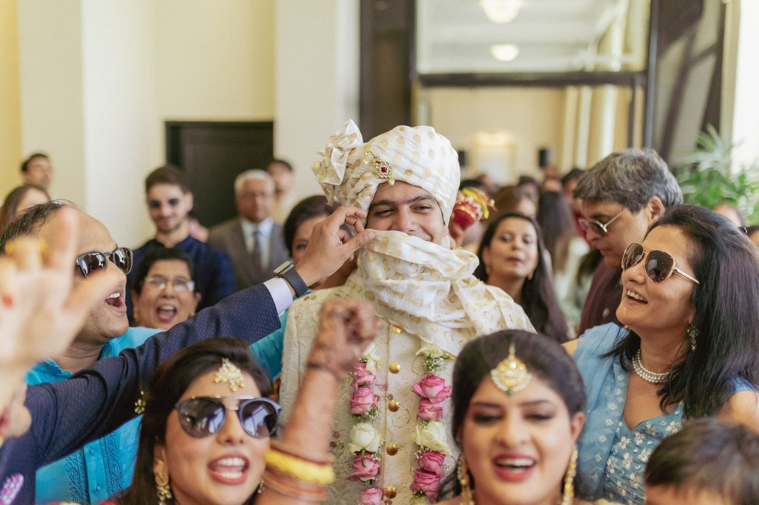 Indian groom in white sherwani and turban surrounded by joyful wedding guests.