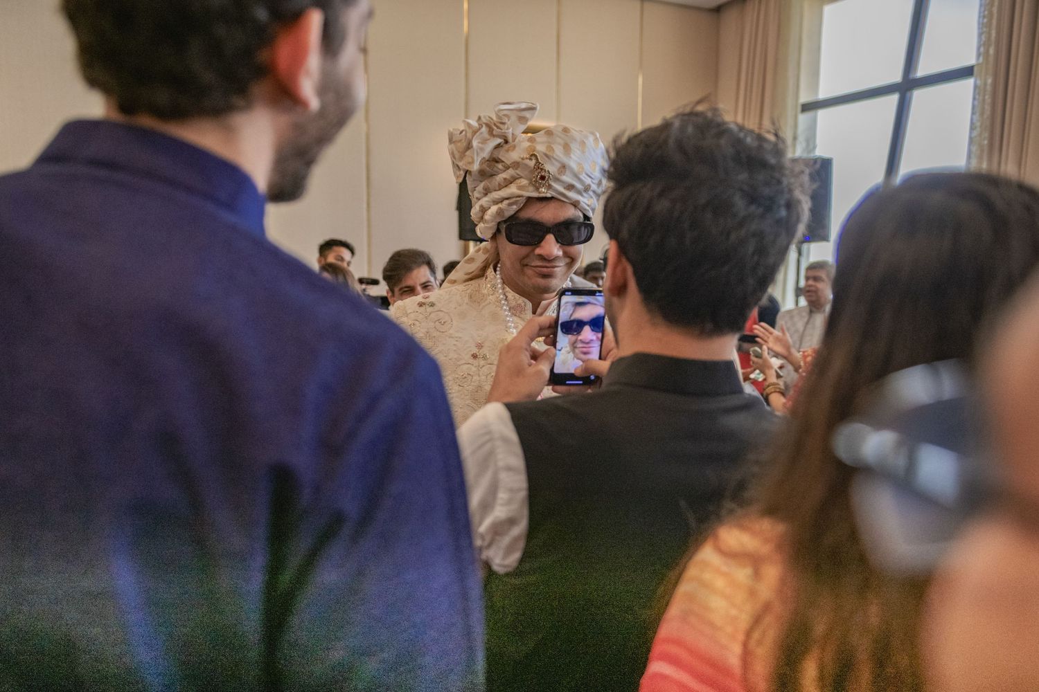 Guest in elaborate gold headdress and sunglasses poses for phone photo at Indian wedding event.