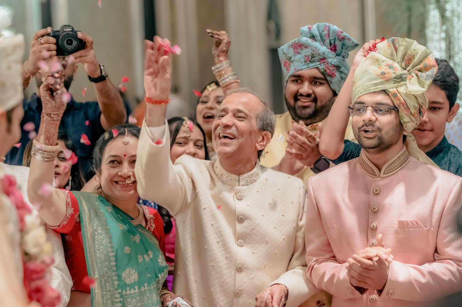 Elderly man celebrates joyfully with raised hands alongside groom in pink sherwani at Indian wedding.