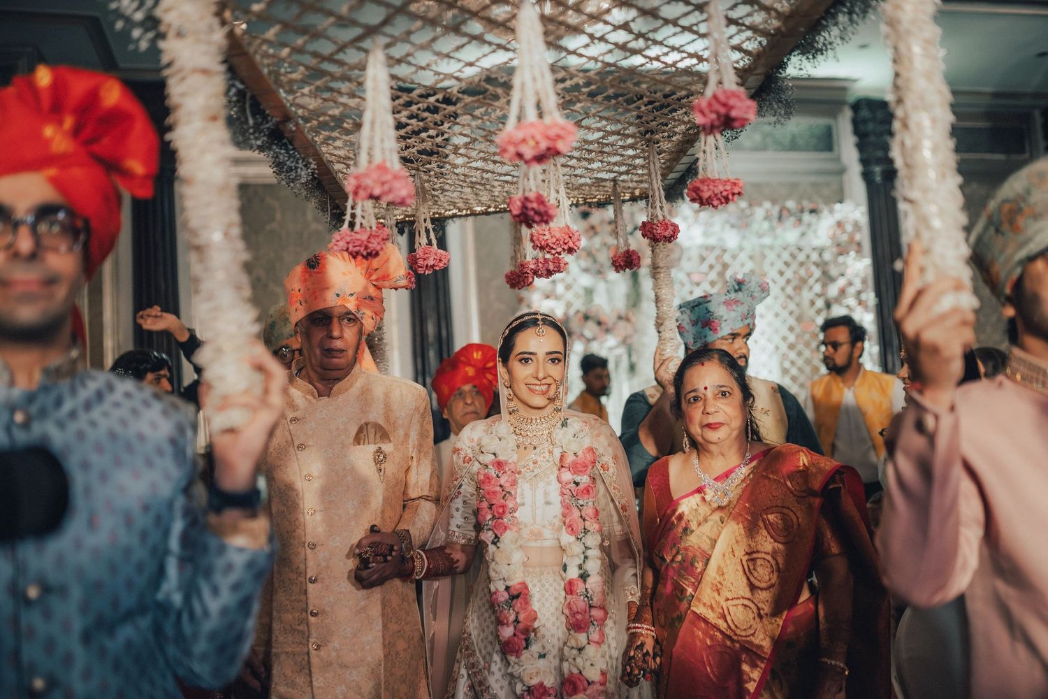 Indian bride walking down the aisle with family under floral canopy at traditional wedding ceremony.