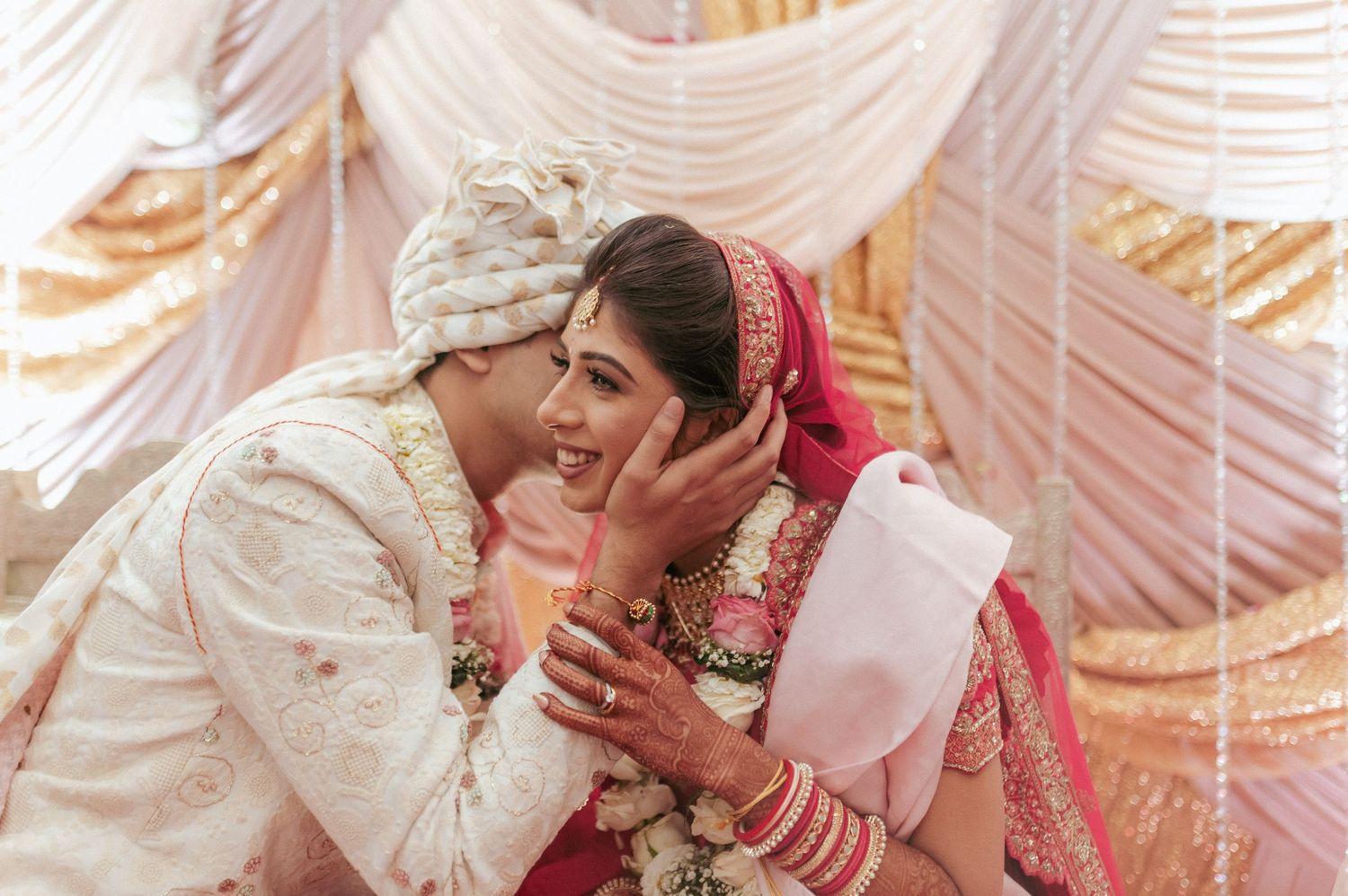 Groom in white sherwani whispers to smiling bride in red lehenga during intimate Hindu wedding ceremony.