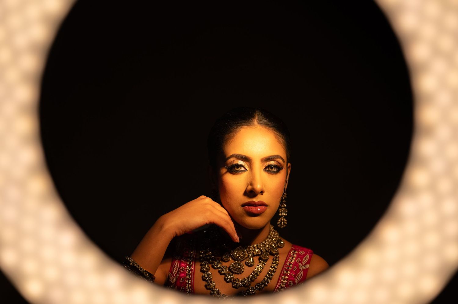 South Asian bride in traditional jewelry framed through circular ring light in dark studio.