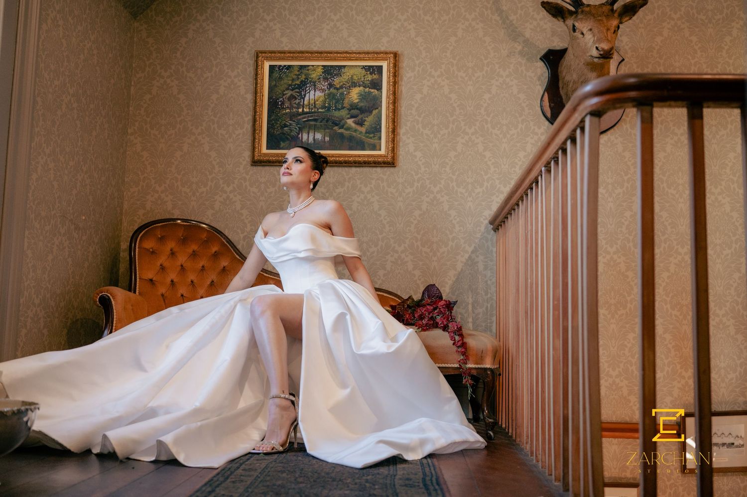 Bride in white off-shoulder ball gown posing gracefully on staircase in vintage decorated room.