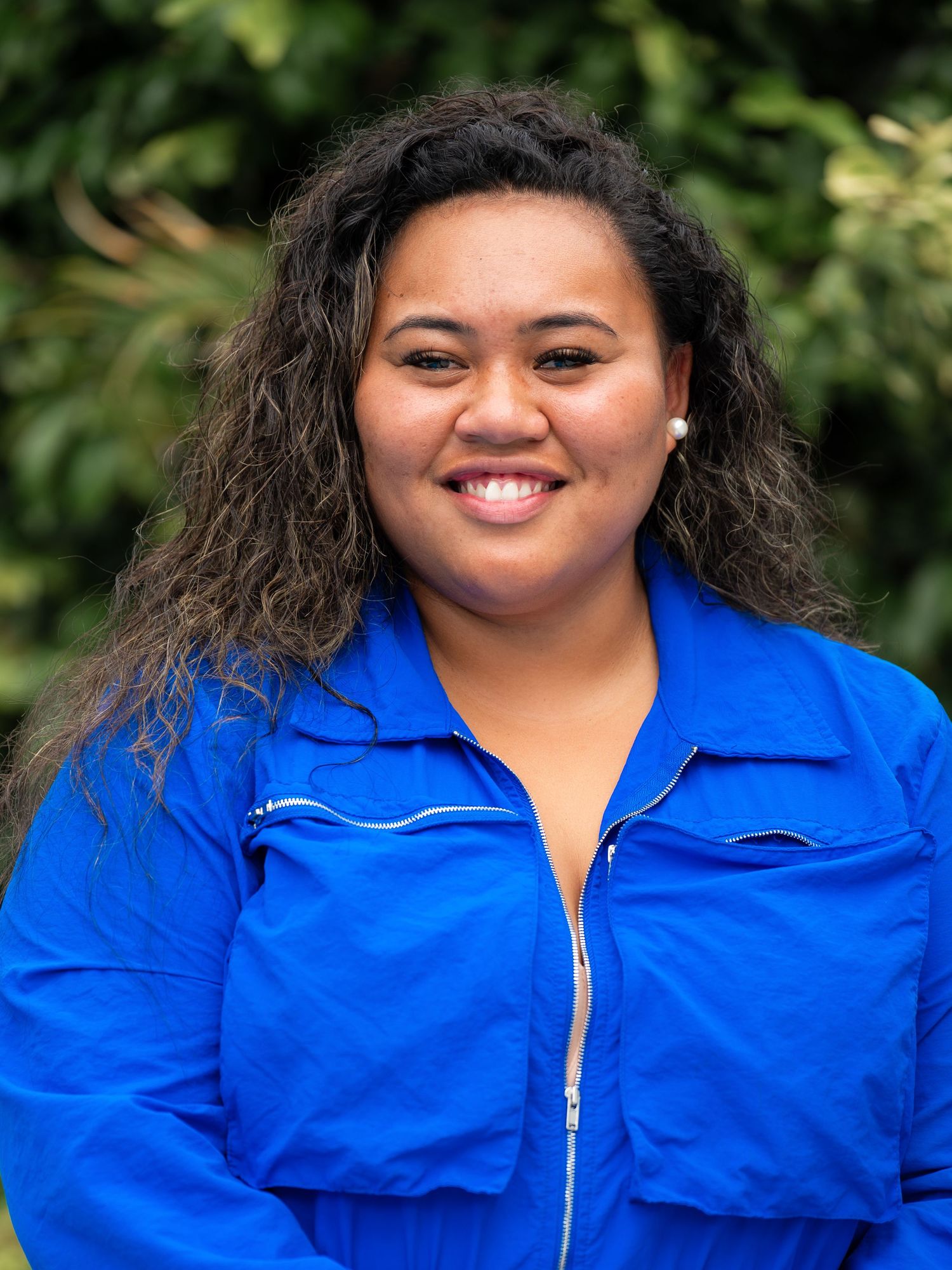 Smiling young woman with curly hair wearing a vibrant blue jacket posed outdoors near green foliage.