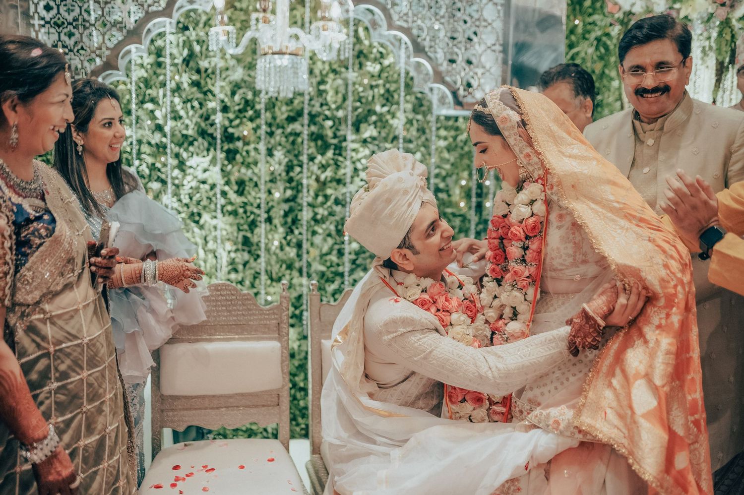 Indian bride and groom exchange garlands while seated at traditional Hindu wedding ceremony with family.