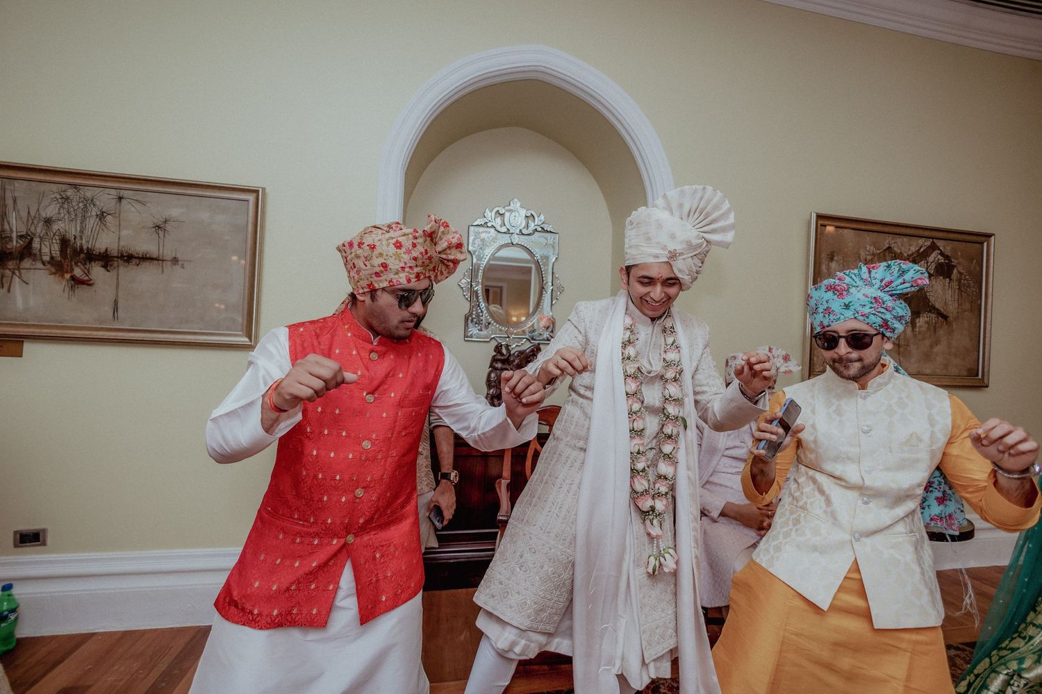 Three men in traditional Indian wedding attire dance together in an elegantly decorated room.