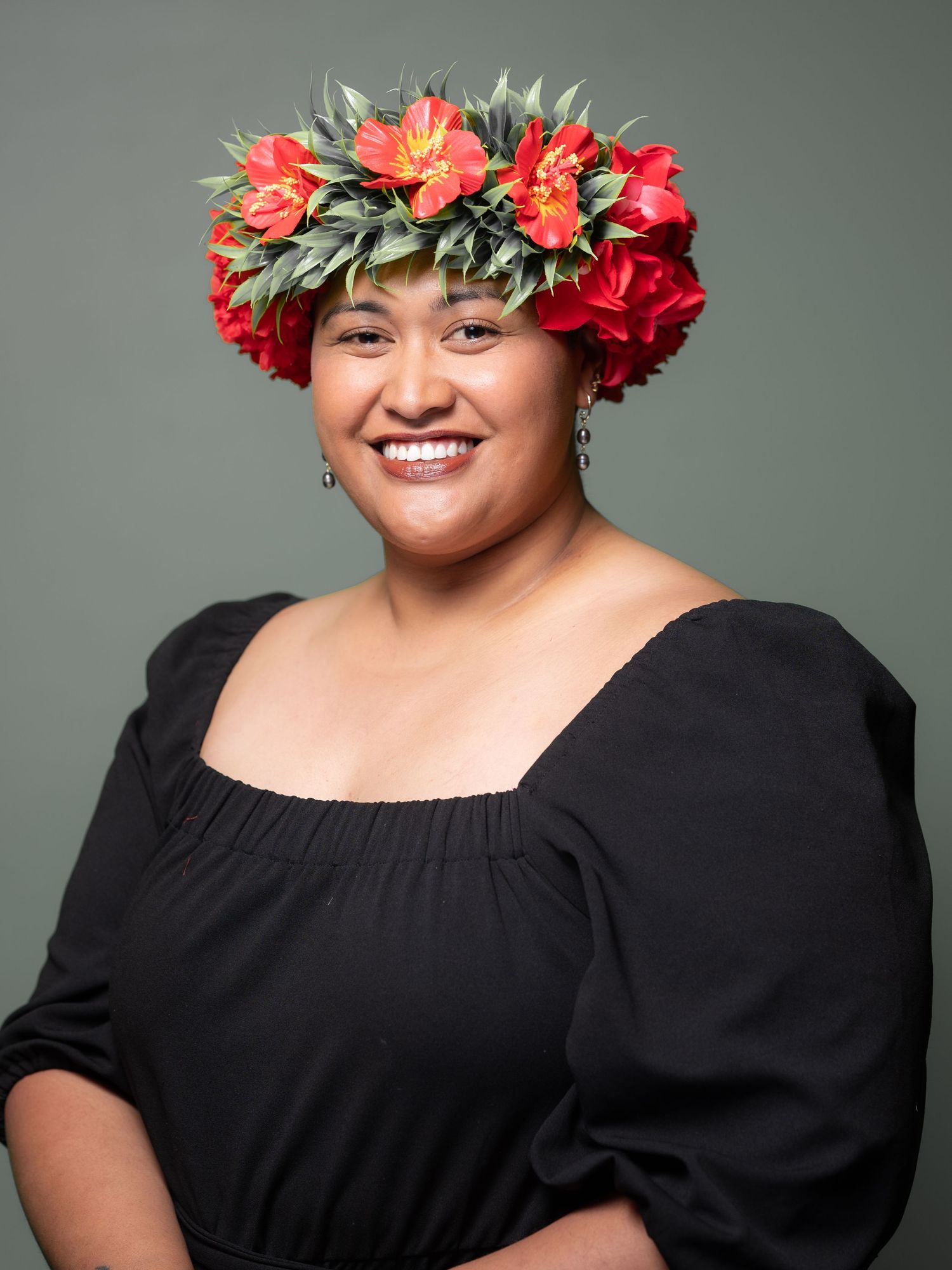 Smiling woman wearing red floral crown and black off-shoulder top against neutral grey background.
