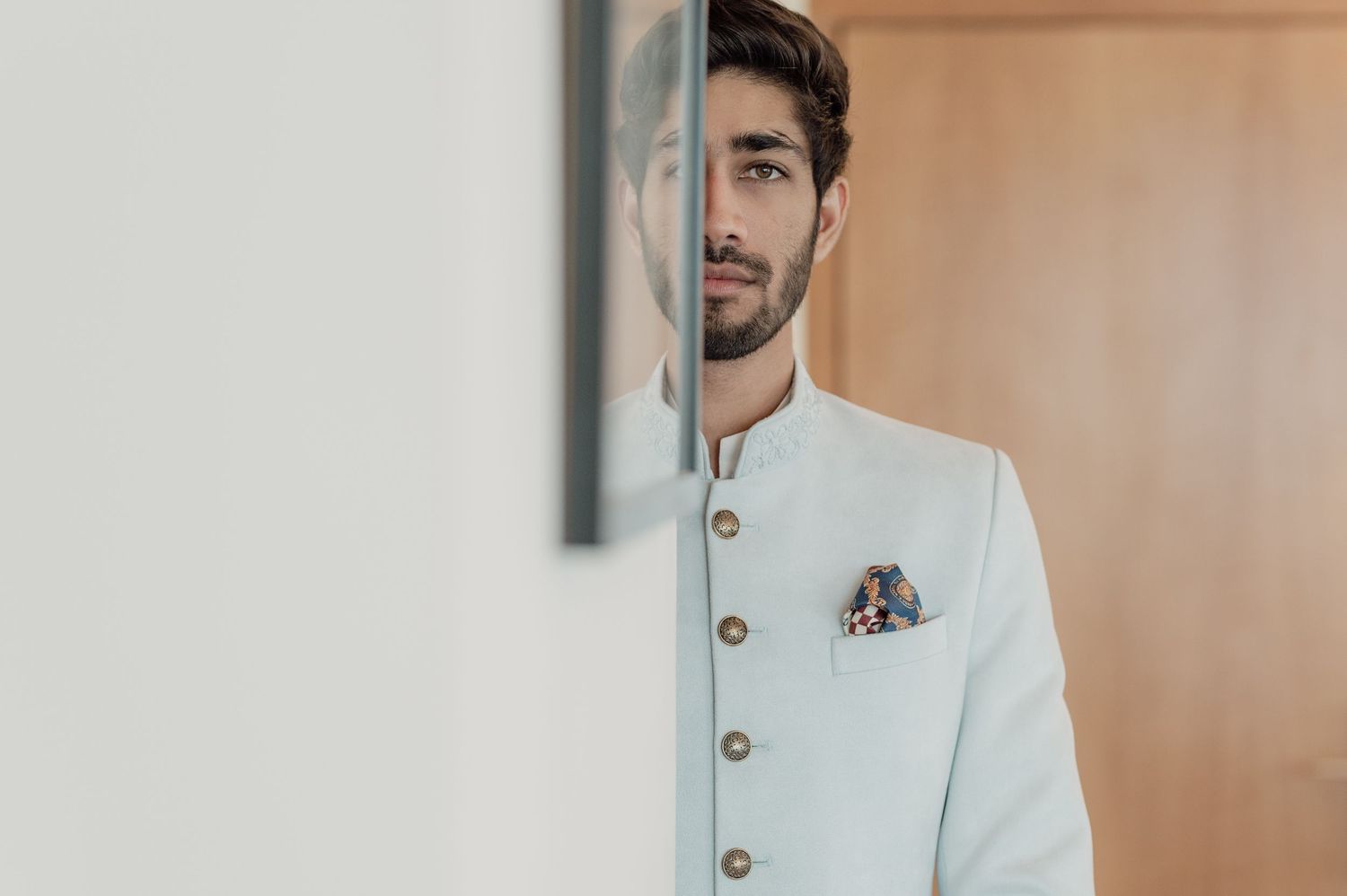 Groom in light blue sherwani with pocket square posing by wall.