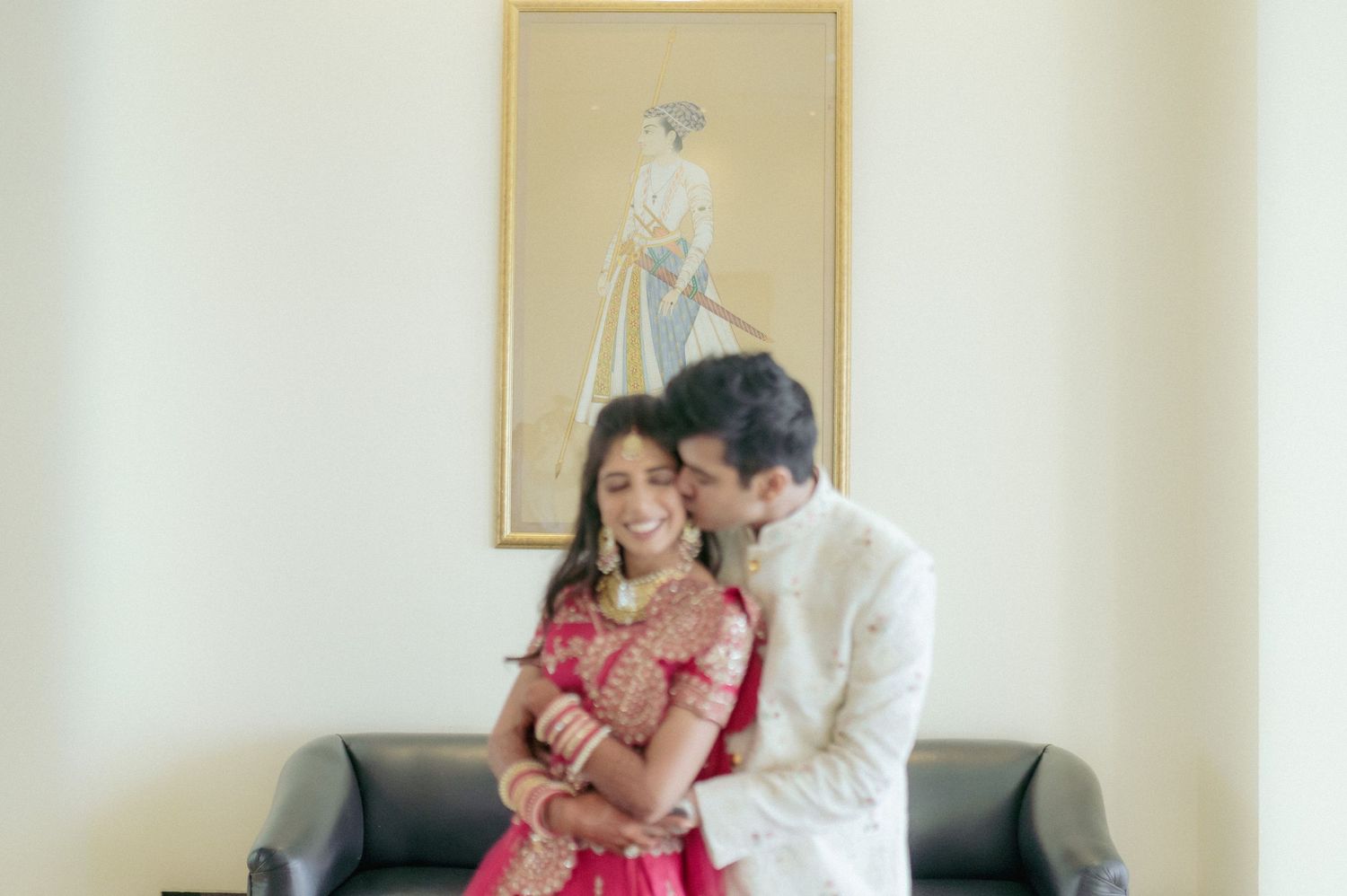 Indian bride and groom sitting together on sofa with traditional artwork hanging behind them.