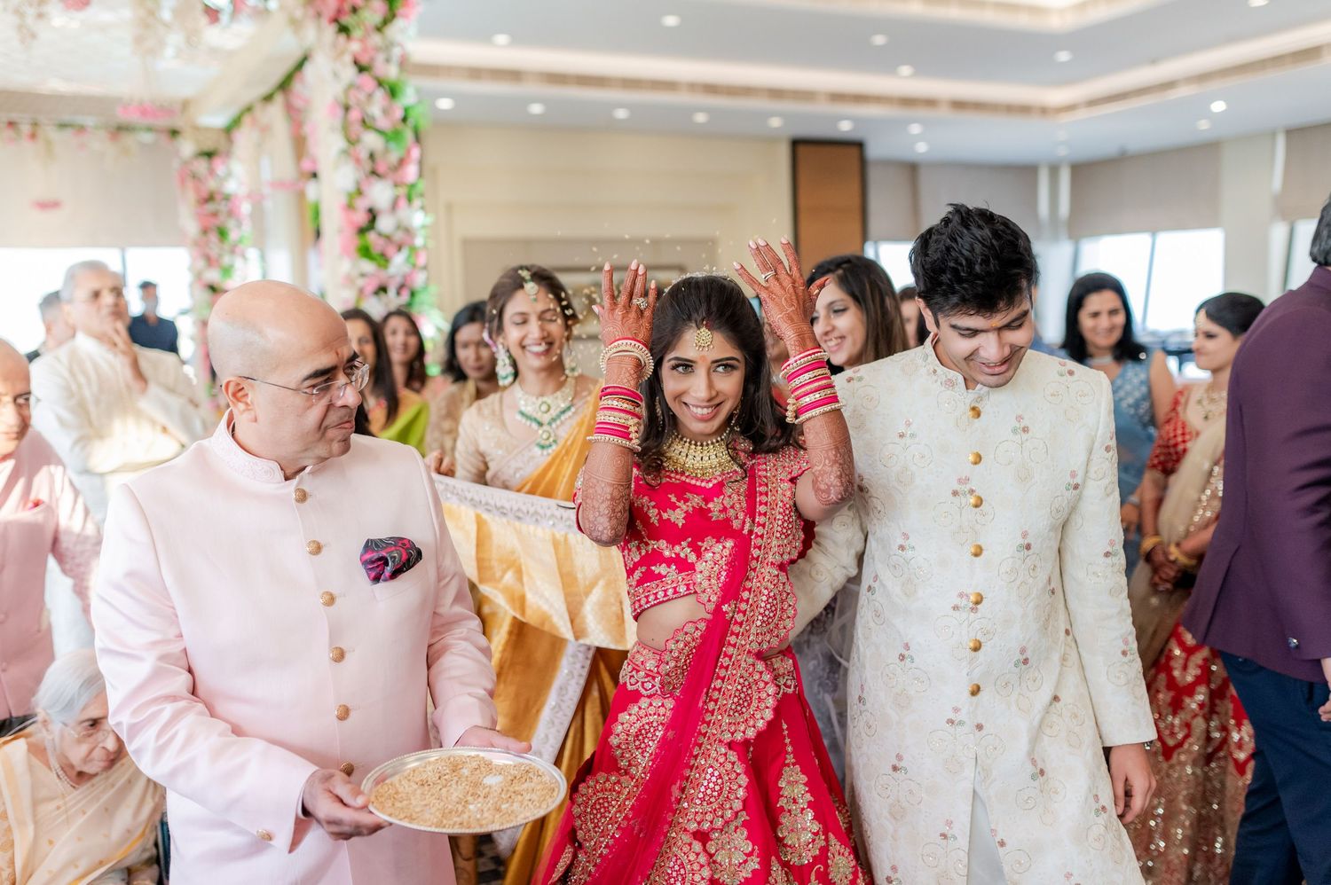 Indian bride in red lehenga walks with groom in white sherwani during traditional wedding ceremony.