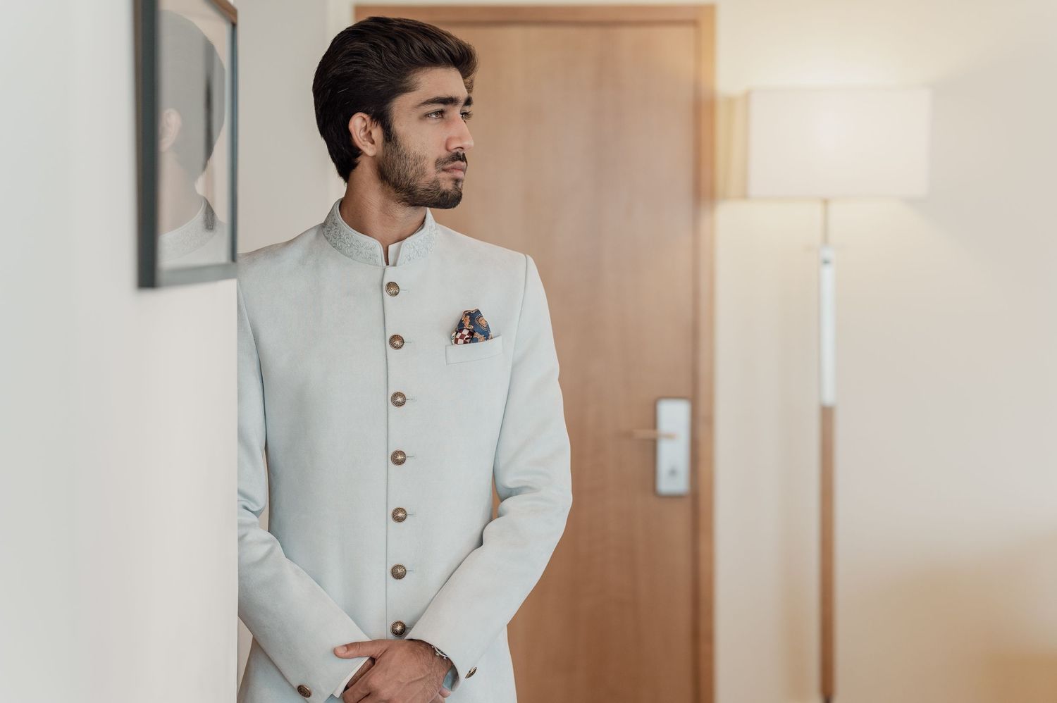 Groom in elegant white bandhgala sherwani standing in hotel room.