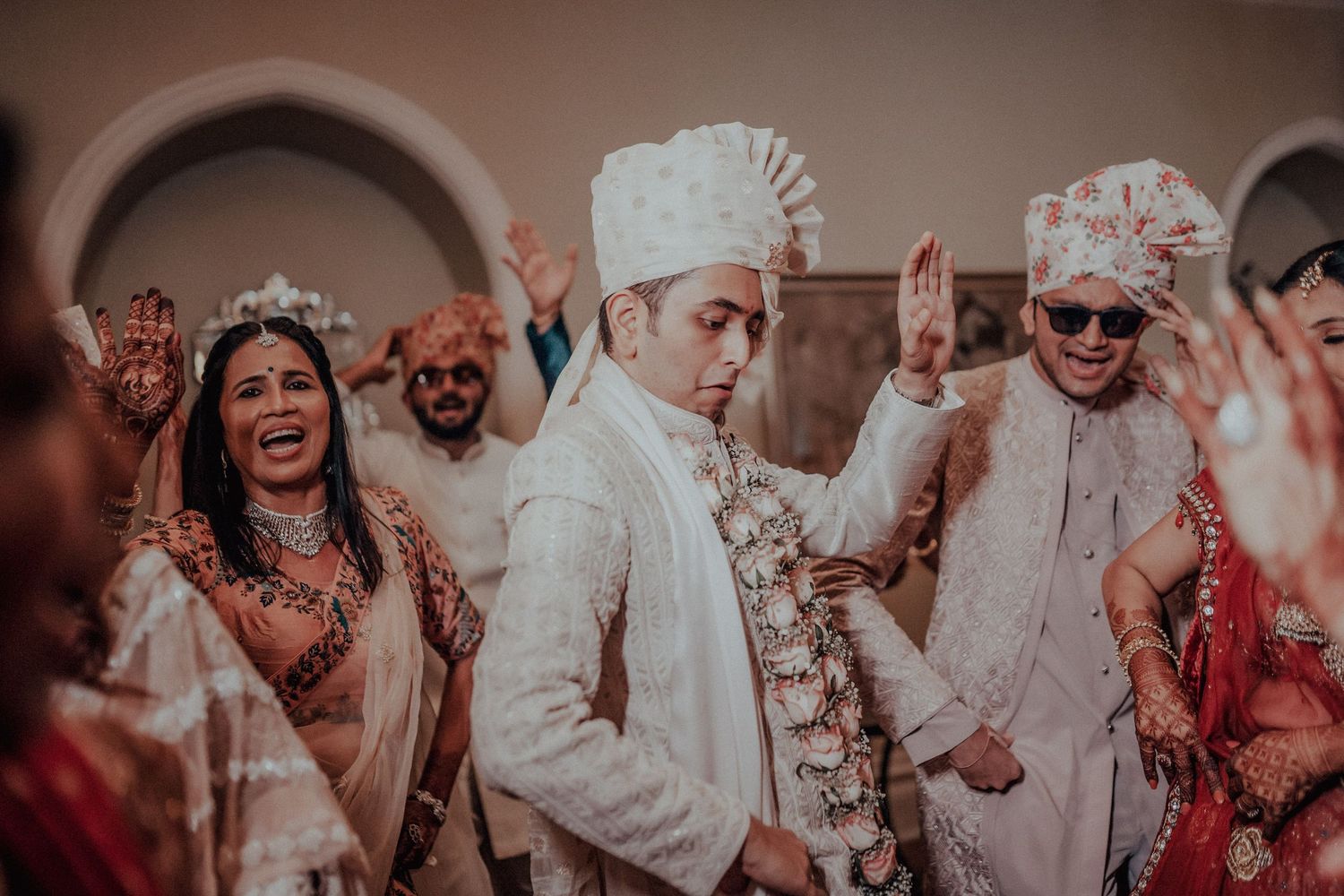Groom in white safa stands between dancing guests at a vibrant Indian wedding celebration.