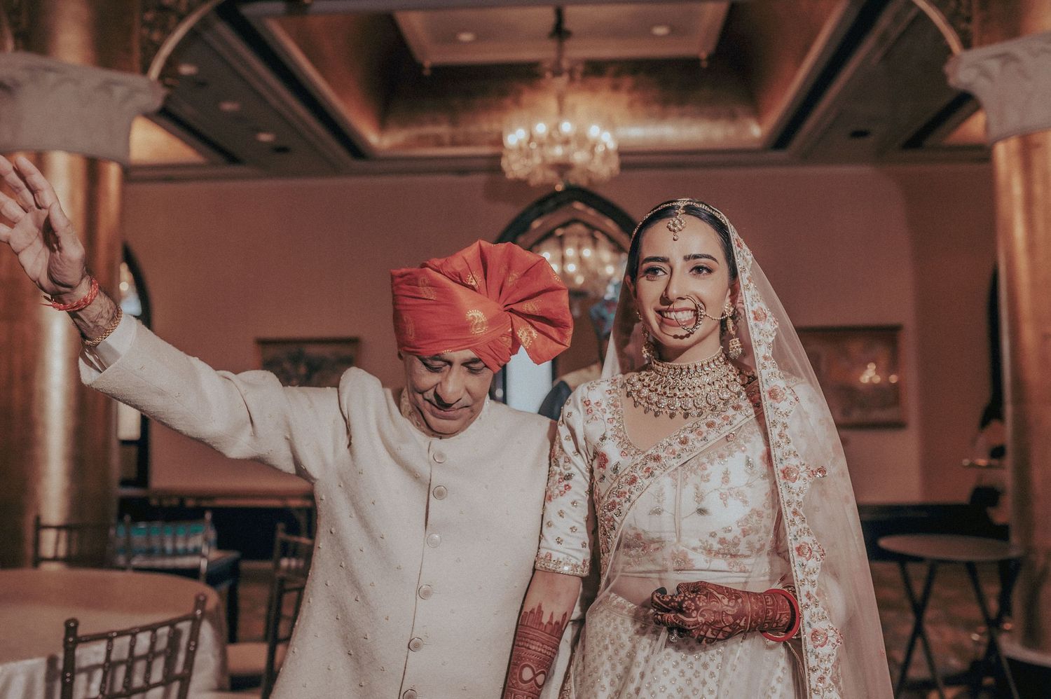 Indian bride in white lehenga smiling joyfully while being escorted by elder in red turban.