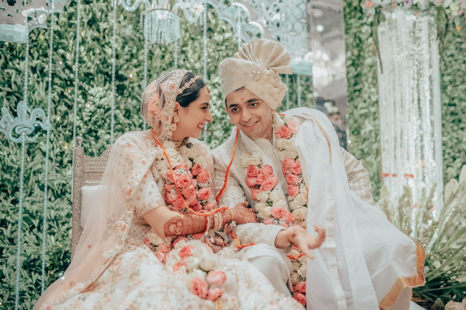 Indian bride and groom in matching white outfits with pink floral garlands smile together.