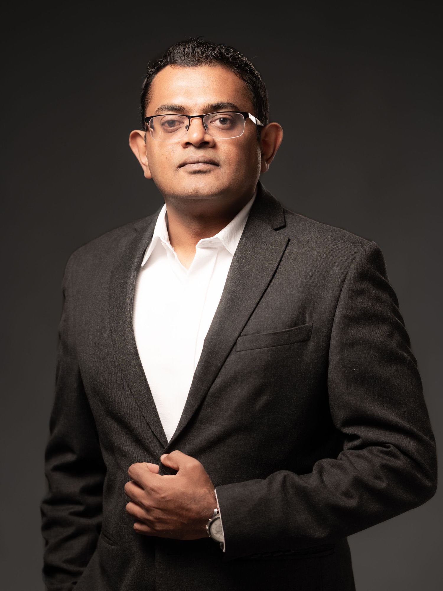 Confident businessman in dark suit and glasses poses for a professional corporate portrait.