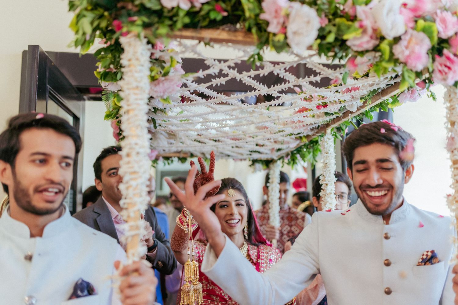 Bride in red lehenga celebrates joyfully under floral mandap with laughing groomsmen at Indian wedding.