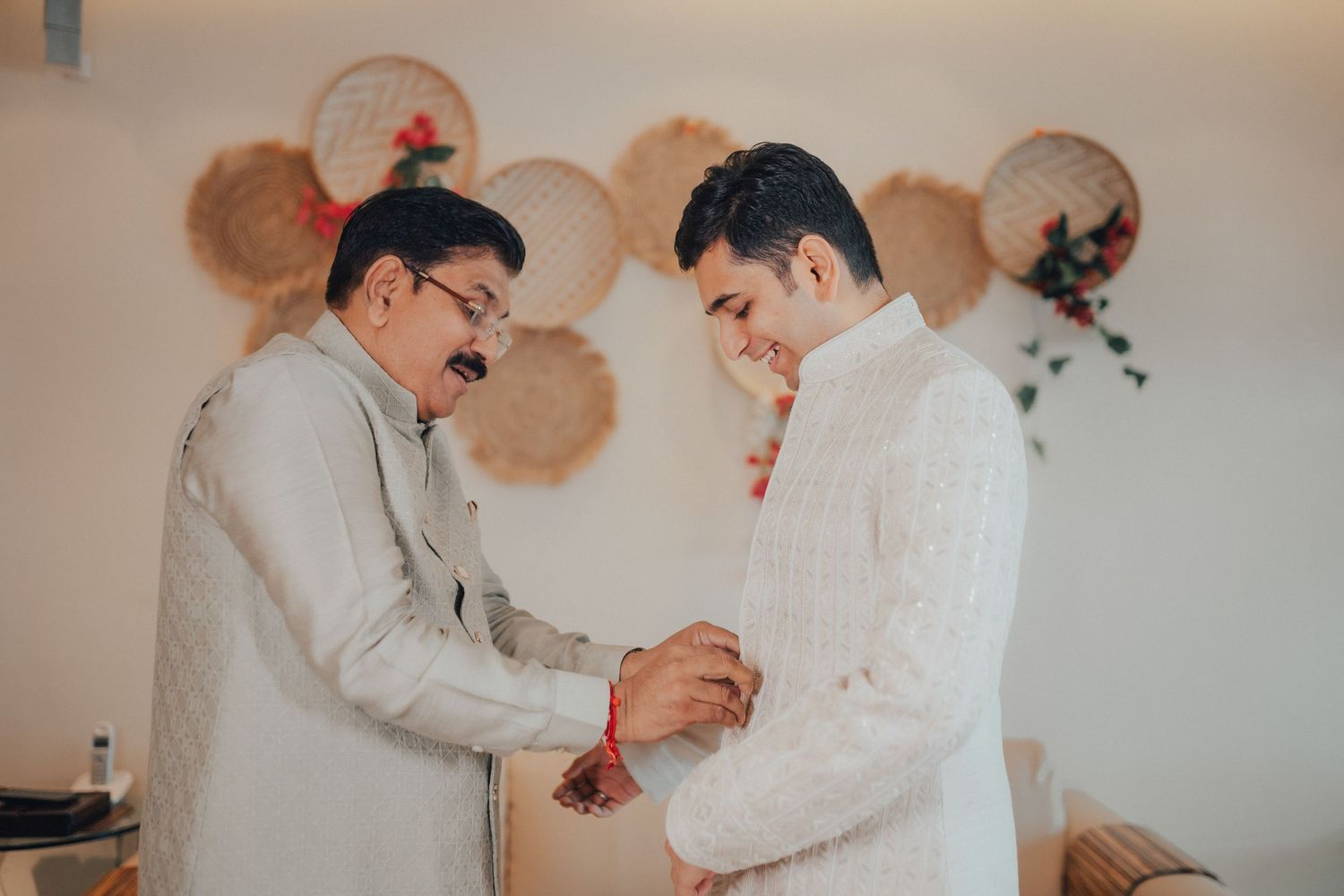 An older man helps a groom in a white sherwani prepare for his Indian wedding ceremony.