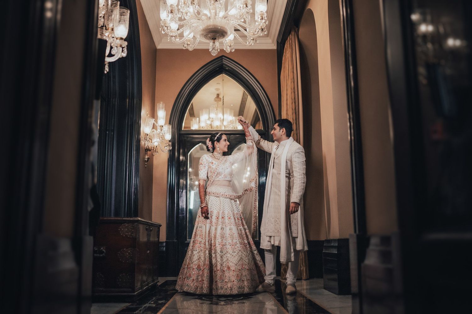 Indian bride in pink lehenga and groom in sherwani pose in grand chandelier-lit palace corridor.