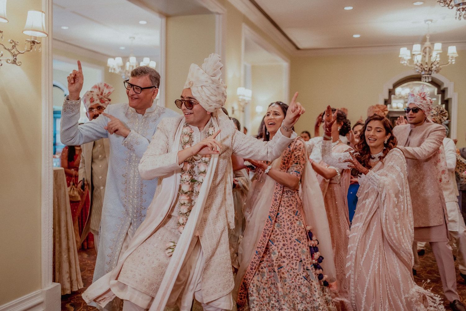 Groom in white sherwani leads a festive procession with family at an Indian wedding.