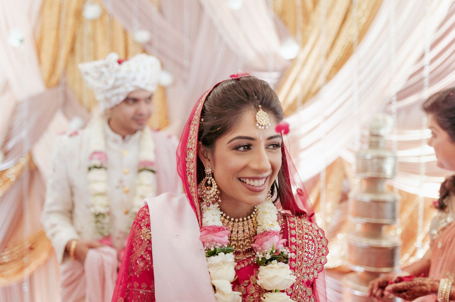 Beautiful Indian bride in red bridal attire adorned with gold jewelry smiles joyfully at wedding.