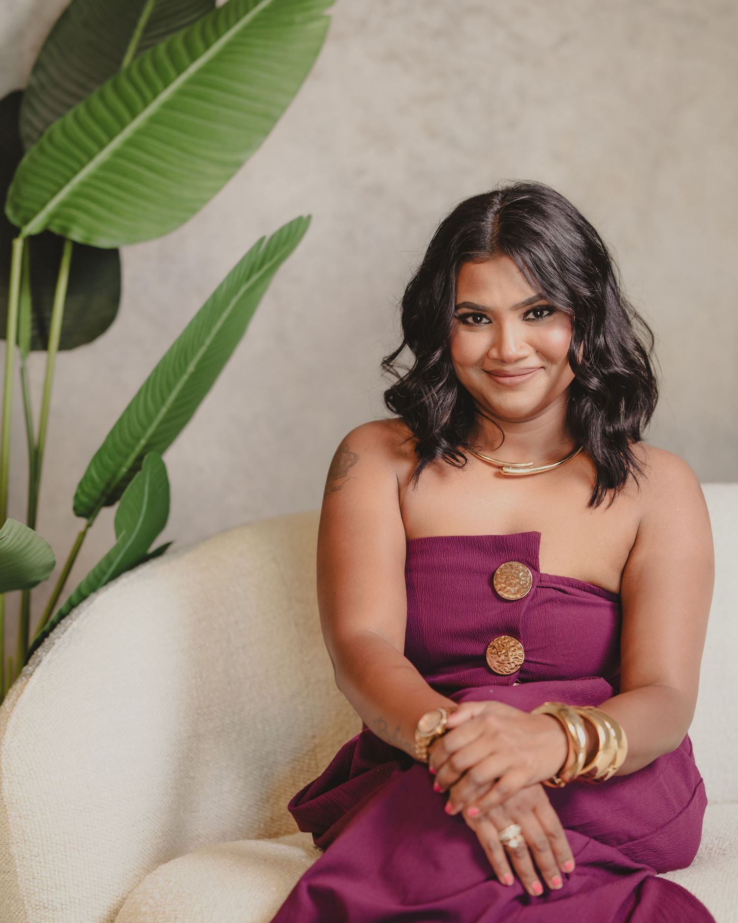 Woman in purple strapless dress with gold jewelry seated in elegant chair near tropical plant.