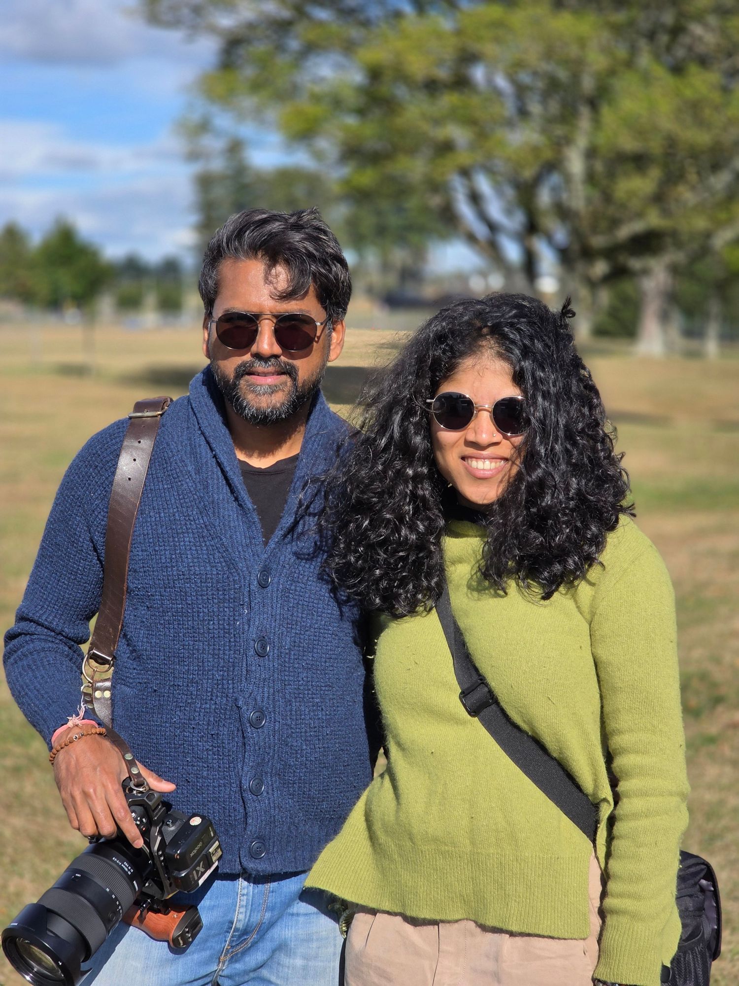 A couple smiling outdoors, man holding a camera, woman with curly hair, in a sunny park.