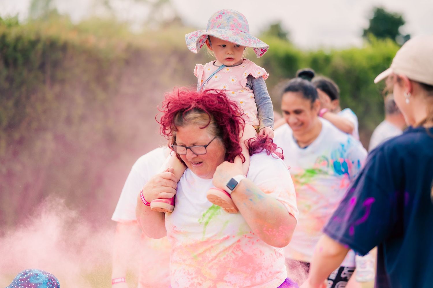 Woman carrying toddler on shoulders both covered in pink powder during outdoor color run event.