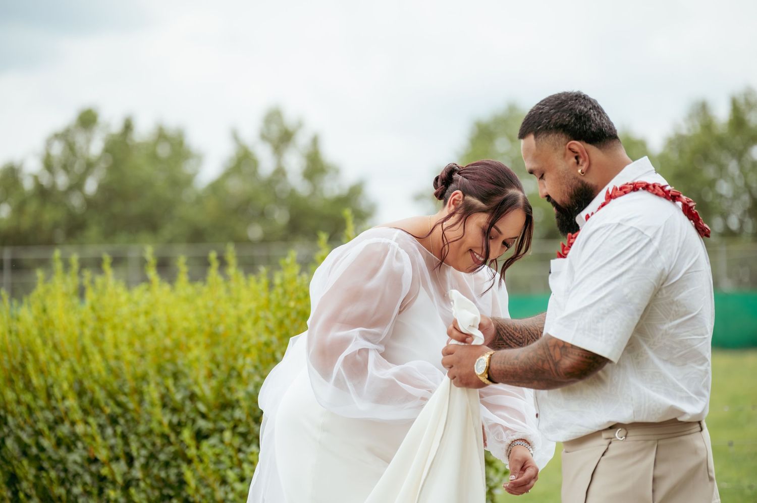 Couple sharing an intimate moment outdoors, groom in white shirt with red lei holding bride's hands.