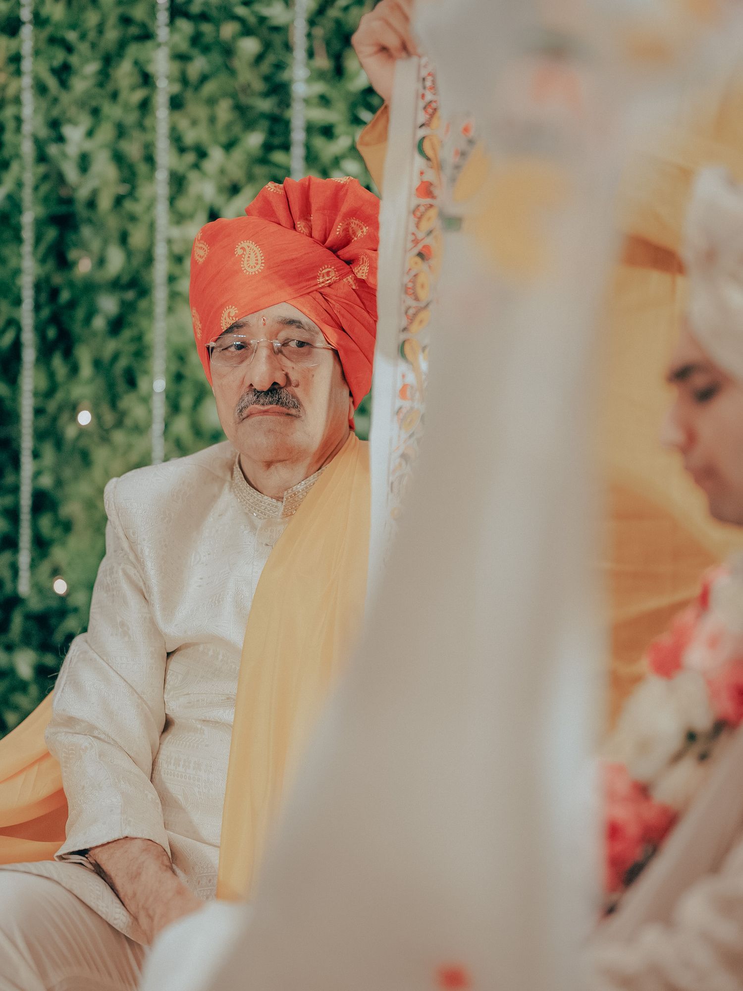 Elder man in orange turban and white sherwani attends traditional Indian wedding ceremony.