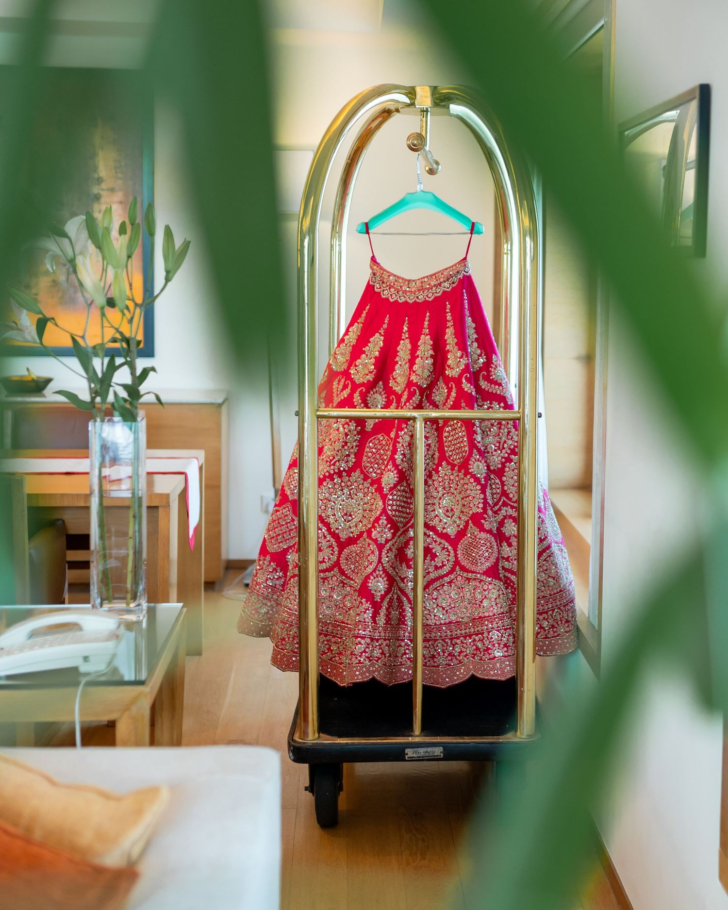 Ornate red and gold Indian bridal lehenga hanging on a luggage cart in a hotel room.