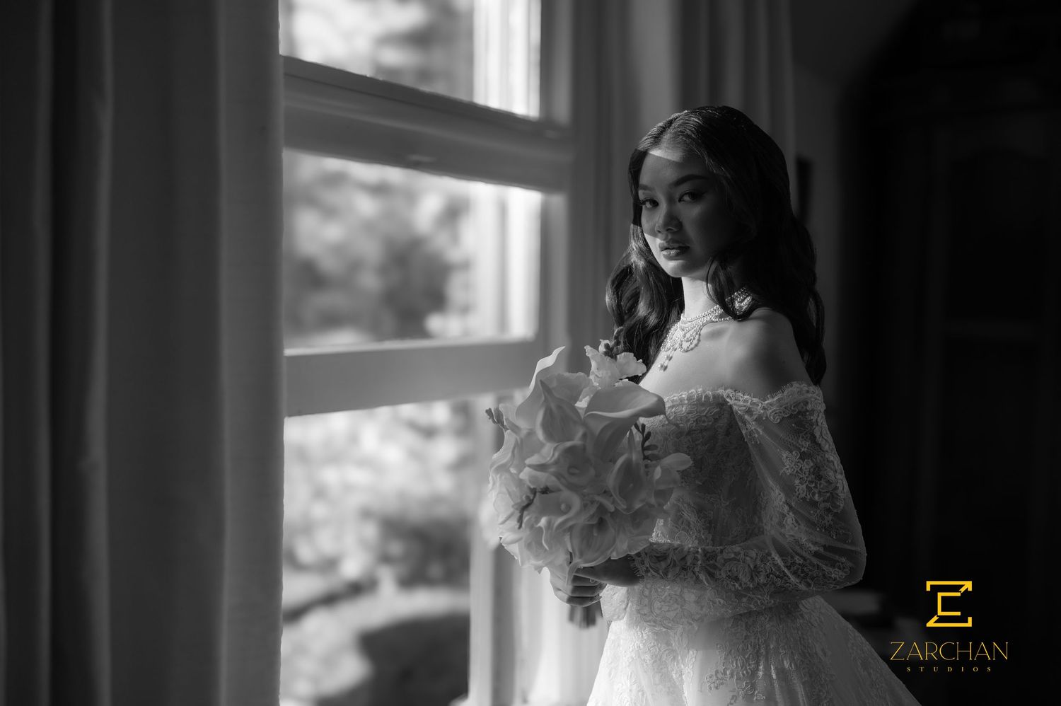 Bride in white lace gown holding bouquet by window in black and white wedding portrait.