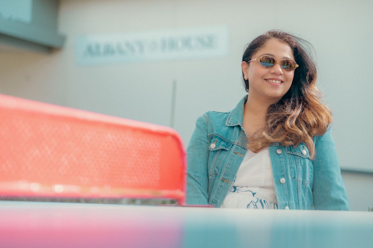 Young woman in denim jacket and sunglasses smiles outdoors near Albany House on a bright day.