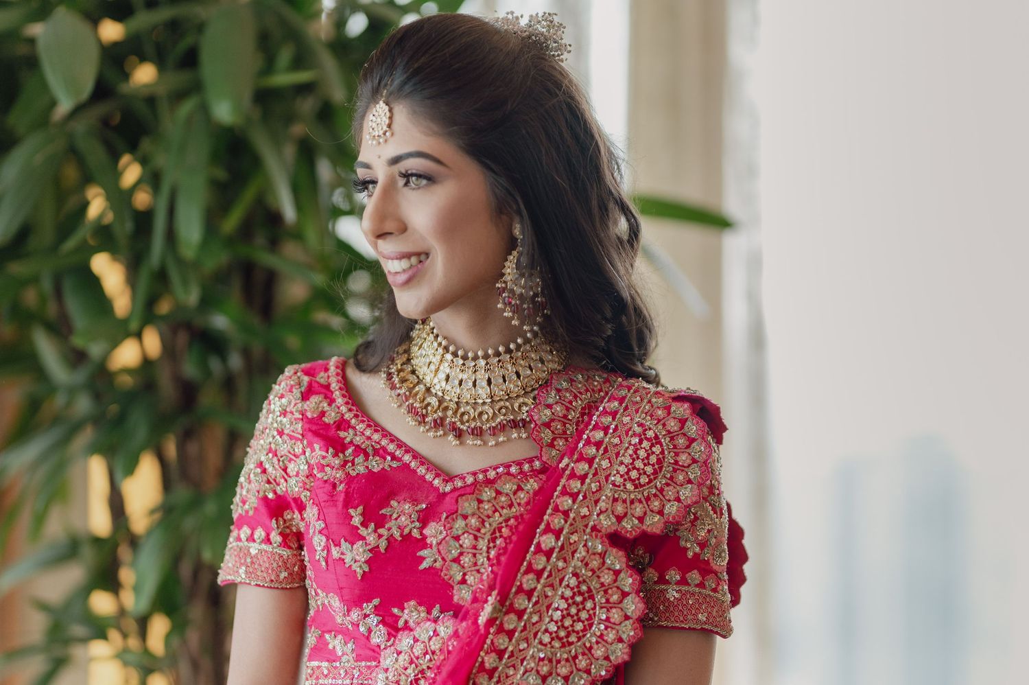 Beautiful Indian bride in pink lehenga with gold kundan jewelry smiling near greenery.