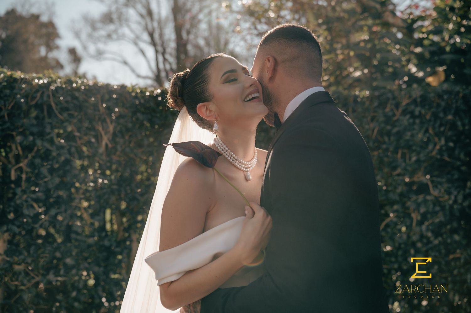 Laughing bride in white gown and veil embraced by groom in suit outdoors in sunlight.