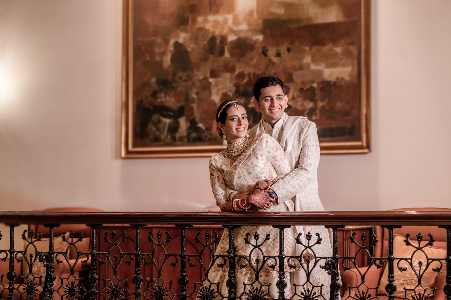 Smiling Indian bride and groom in traditional white attire pose on ornate iron balcony railing.