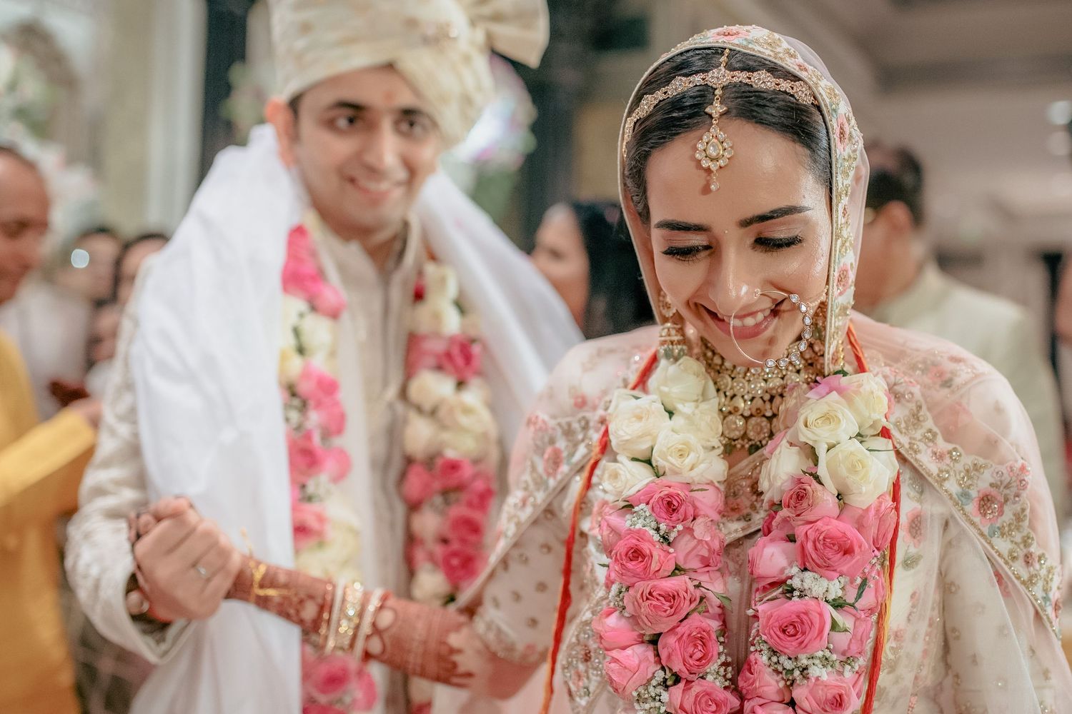 Smiling Indian bride adorned with floral garland and gold jewelry holds grooms hand at wedding ceremony.