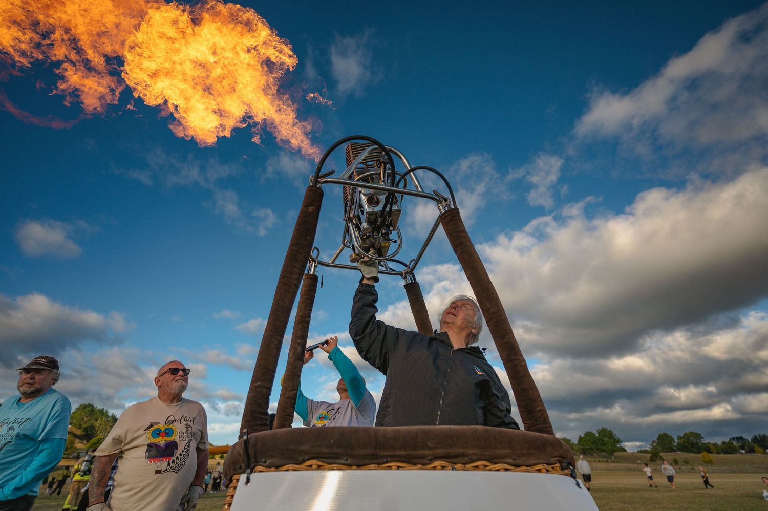 Man operates hot air balloon burner producing large flame against blue cloudy sky at outdoor event.