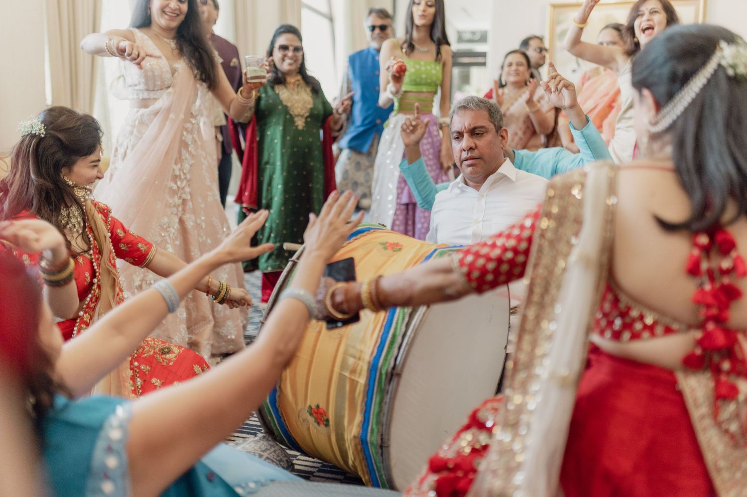 Energetic Indian wedding guests dancing around dhol drummer in colorful traditional attire.