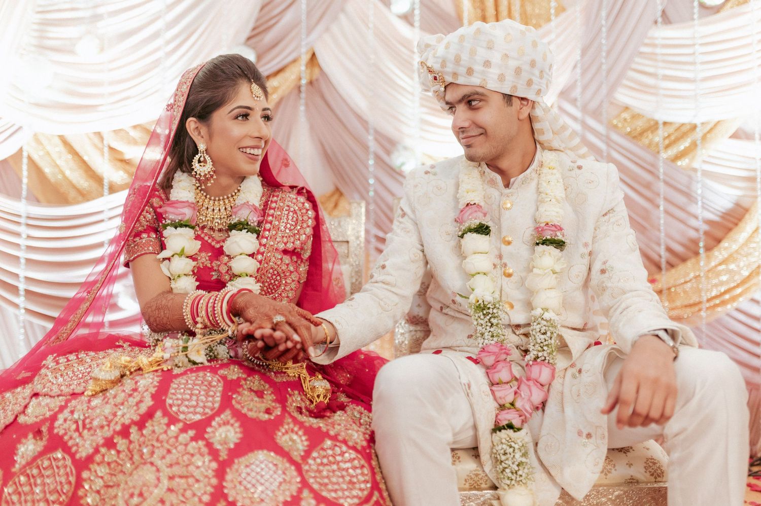 Indian bride and groom in traditional red and white wedding attire smiling during ceremony.