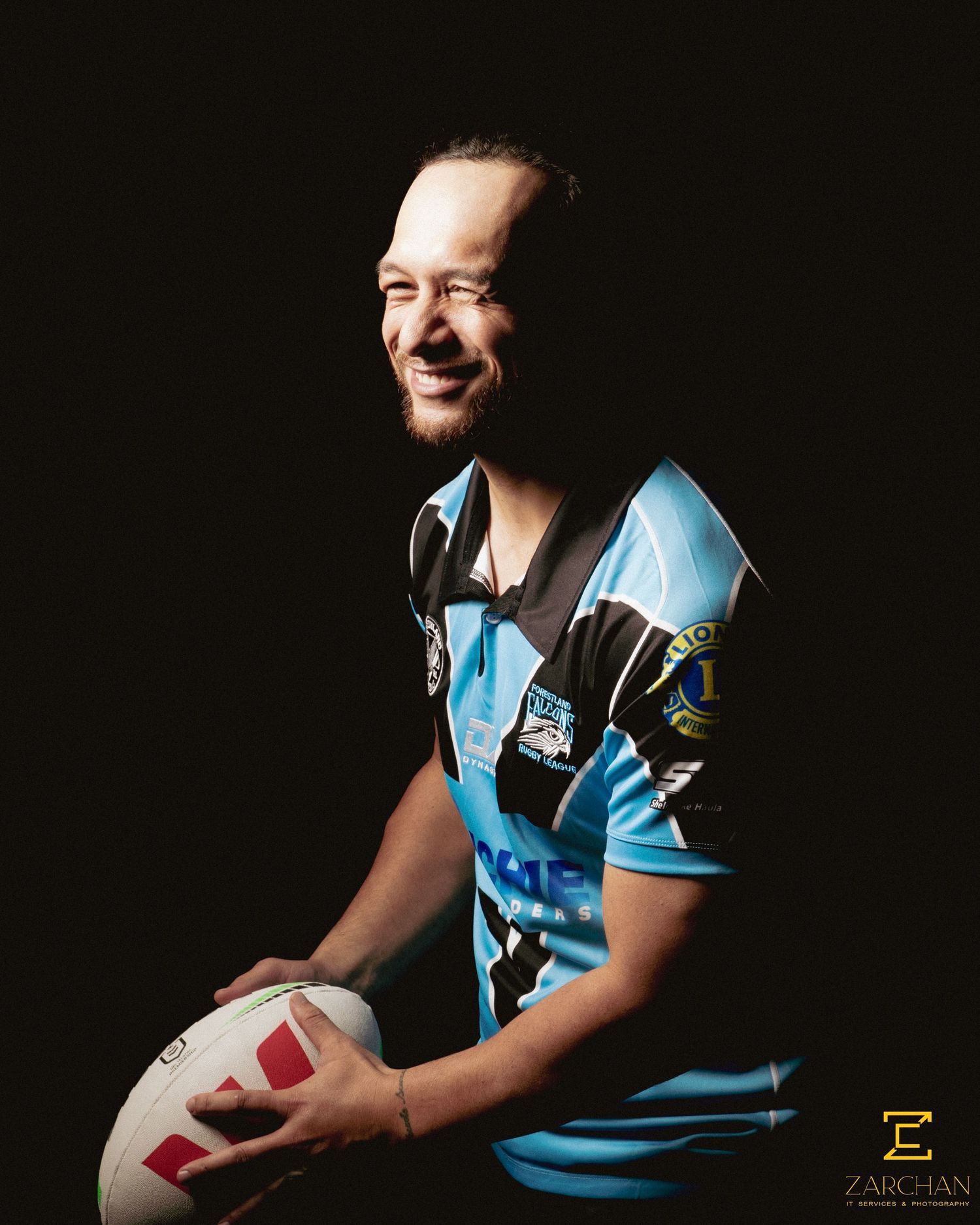 Smiling rugby league player in blue and black jersey holds a football in a dark studio.