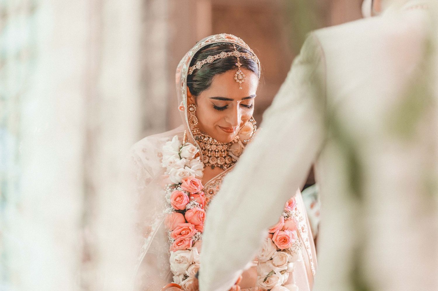 Beautiful Indian bride in white lehenga wearing traditional gold jewelry and floral garland at wedding.