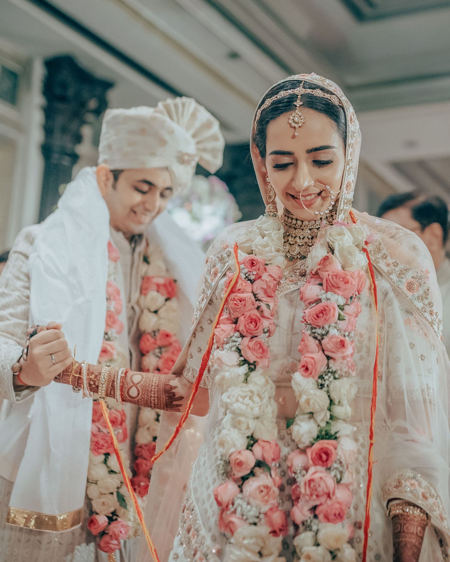 Indian bride in embroidered lehenga with pink rose garland holds red thread during traditional wedding ritual.